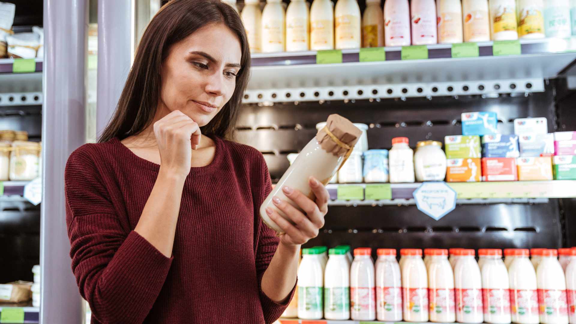 A young woman with brown hair, looks at a bottle of milk. She appears to be reading the ingredients. She stands in a supermarket isle by the milk and dairy products.